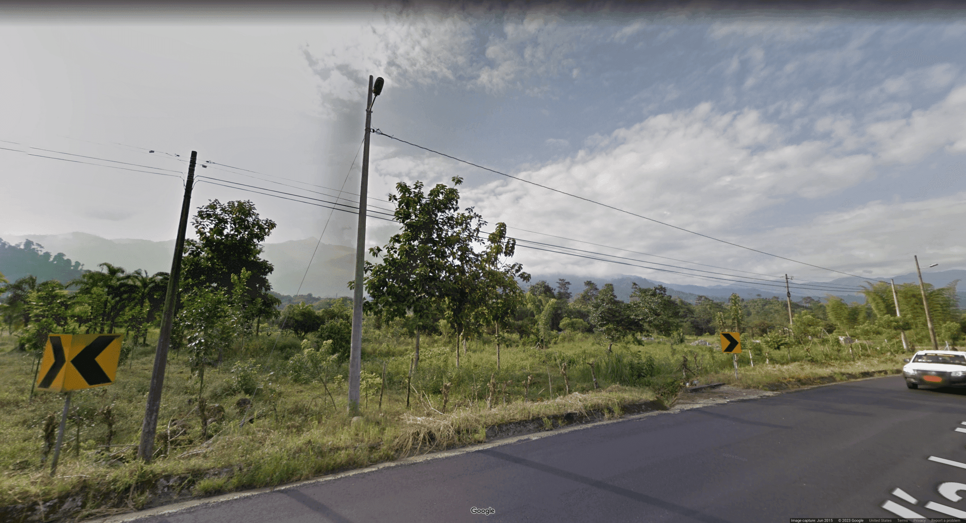 Picture of a roadside in a seemingly tropical area. There are black on yellow turn chevrons on the edges of the road, and a car off to the right with an orange license plate